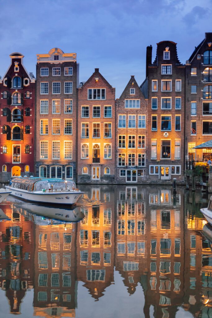 Amsterdamse gracht met herenhuizen en een rondvaartboot in het avondlicht.
