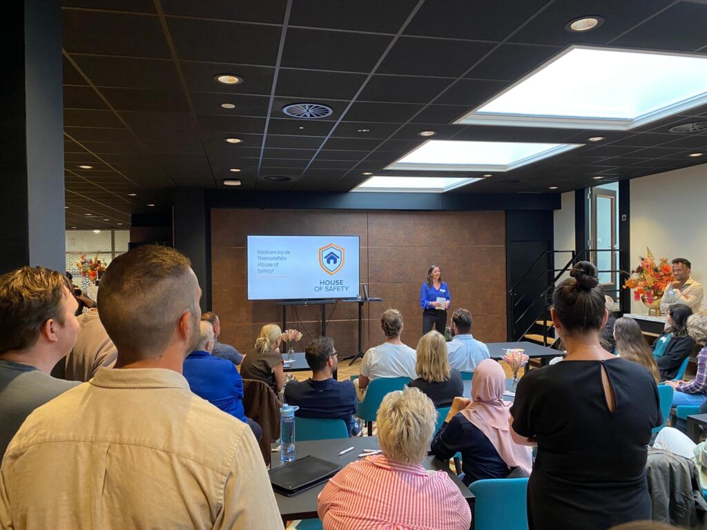 Foto van de zaal waar de bijeenkomst plaatsvindt. Hierop is de spreker en zijn de aanweizigen te zien.